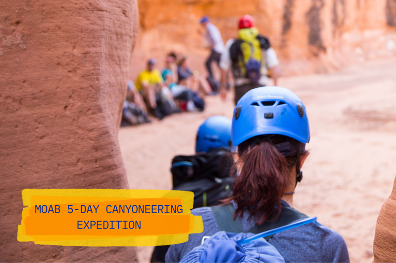 The image shows a group of people participating in a 5-day canyoneering expedition in Moab. They are walking through a narrow canyon, wearing helmets and backpacks, suggesting they are prepared for outdoor activities. The focus is on the person in the front with a blue helmet, with the rest of the group blurred in the background.